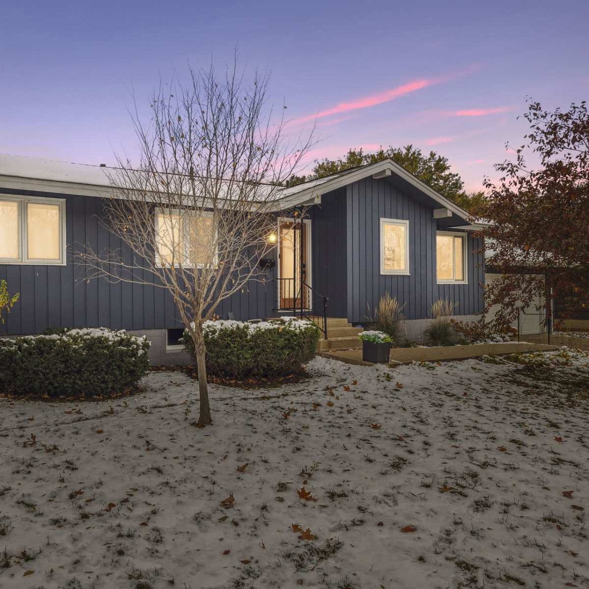 A blue single-story house with white accents is surrounded by a snow-covered yard and bare trees at dusk.