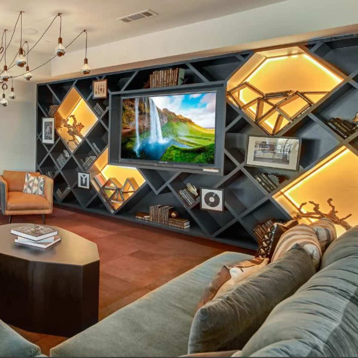 A modern living room featuring a sectional sofa, a coffee table, and a wall-mounted television set against a geometric bookshelf with decorative lighting.