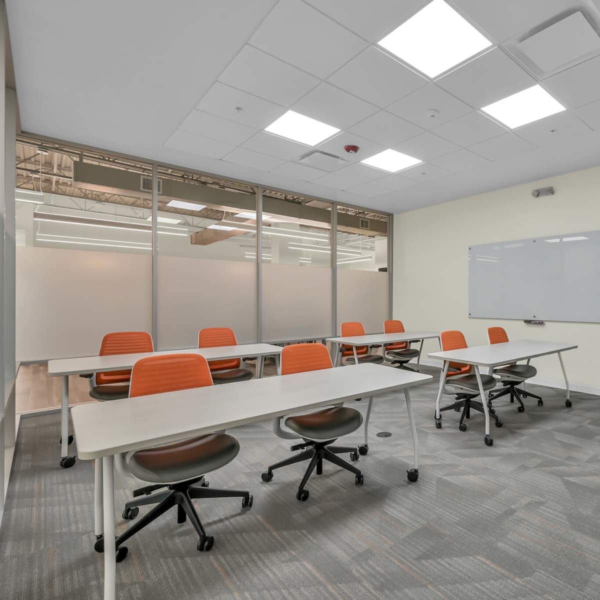 A modern classroom features several tables with orange chairs, a whiteboard, and large windows along one wall.