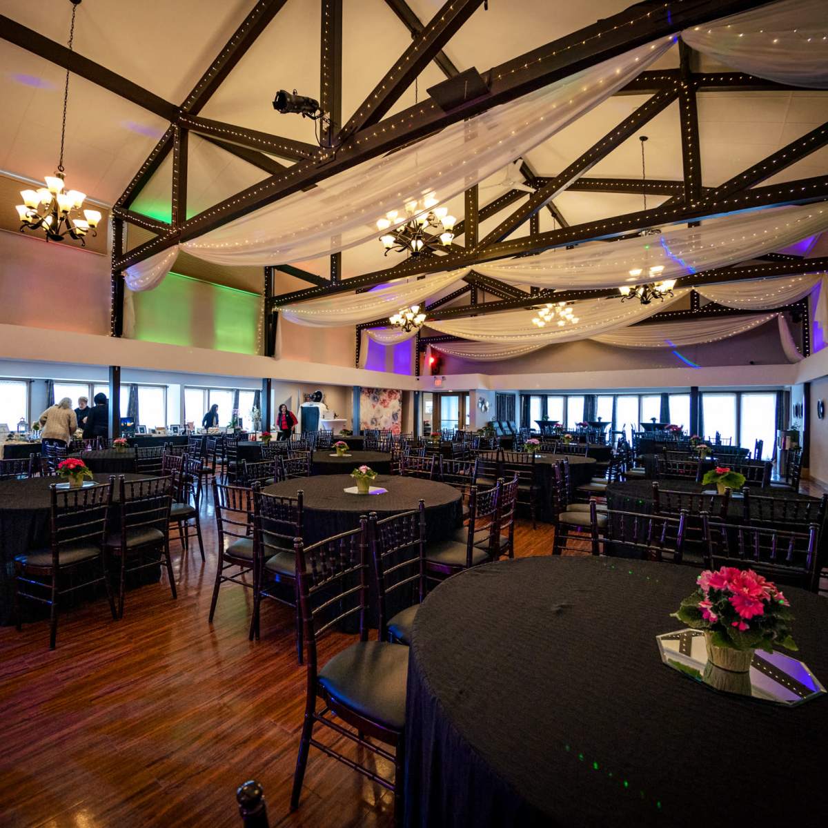 An elegantly arranged banquet hall with black tables, chairs, and floral centerpieces, featuring high ceilings and decorative lighting.