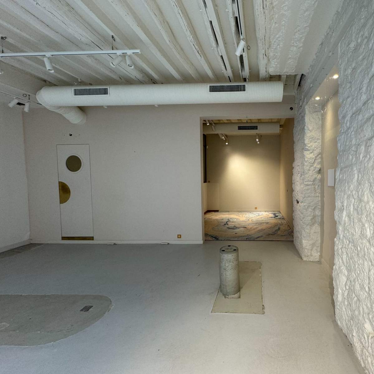 An empty room with a textured white wall, a circular doorway, and an adjoining room illuminated by overhead lights.