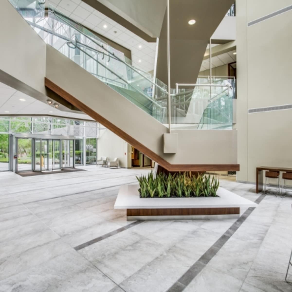 A modern lobby with a staircase, glass walls, and a central planter surrounded by gray tiles.