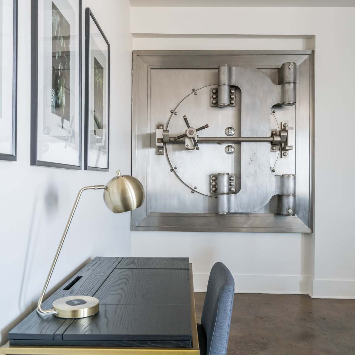 A sleek black desk with a brass desk lamp is positioned in front of a large, metallic safe embedded in the wall.