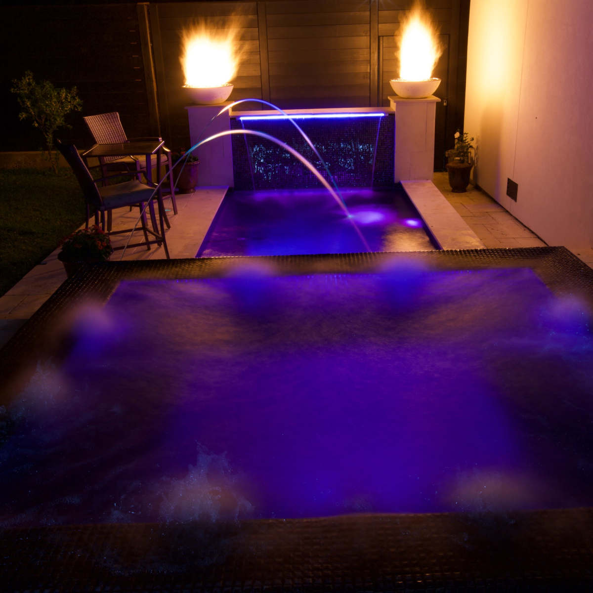 A backyard area with two illuminated water features, one with a fountain and the other with jets, surrounded by a stone deck and a chair.