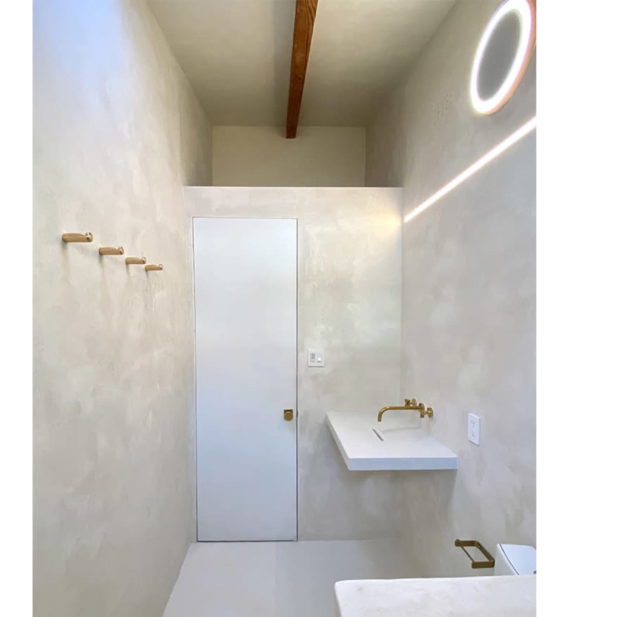 A minimalist bathroom with a white wall, a wooden beam ceiling, a door, a sink with a gold faucet, and several wooden hooks mounted on the wall.