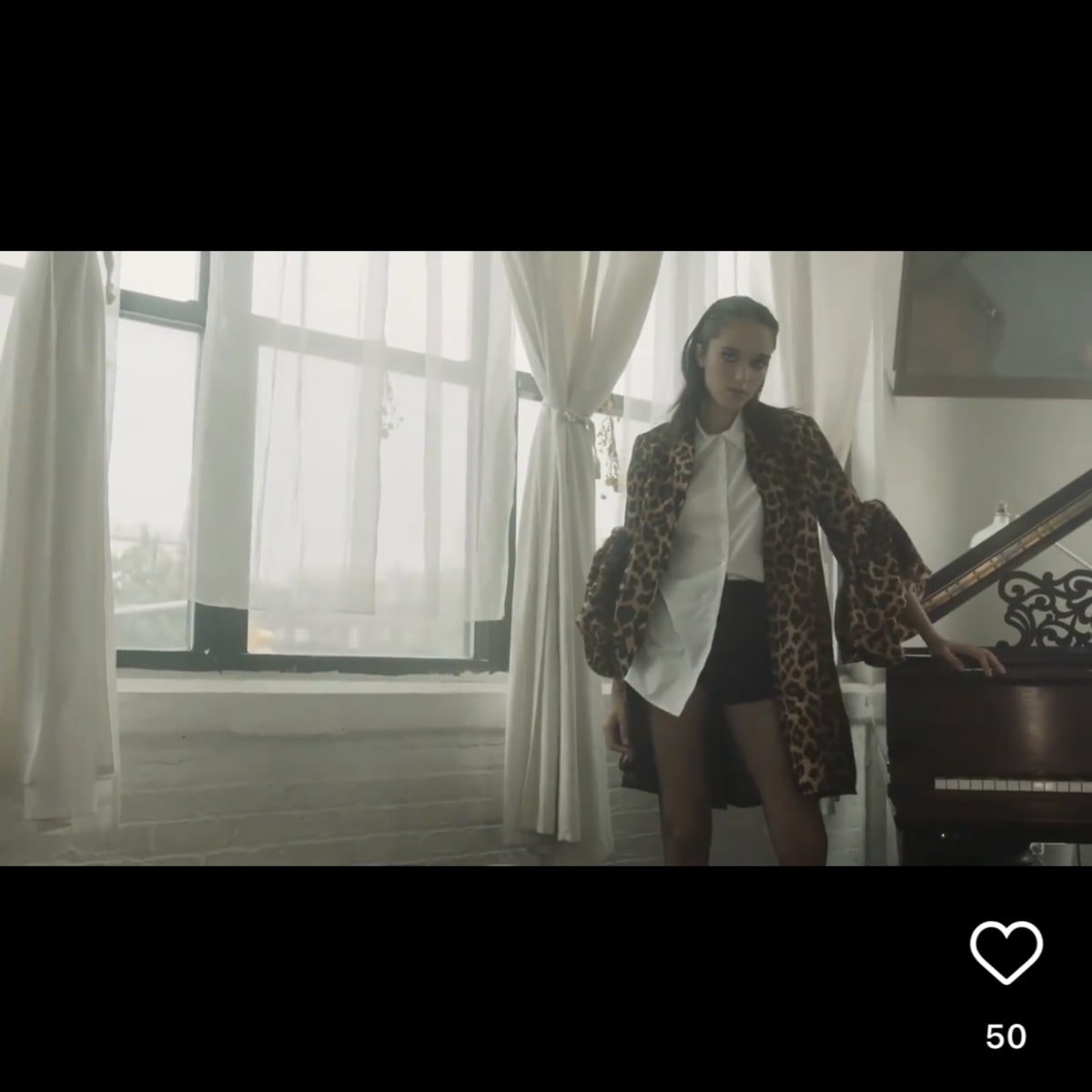 A person stands next to a grand piano in a room with large windows and light-colored drapes.