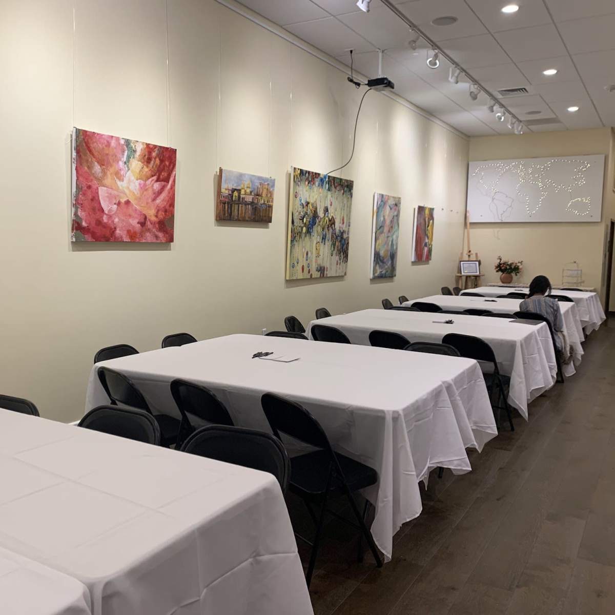 A room with several round tables covered in white tablecloths is lined with colorful abstract paintings on the walls.