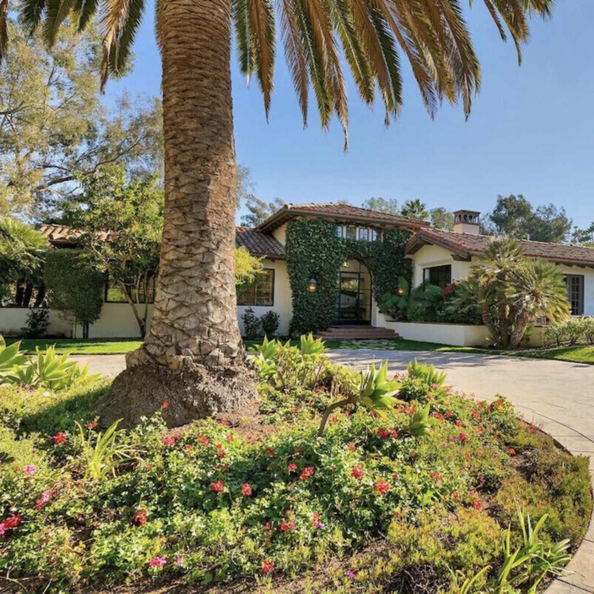 A Mediterranean-style home with a tile roof is surrounded by lush landscaping and palm trees.