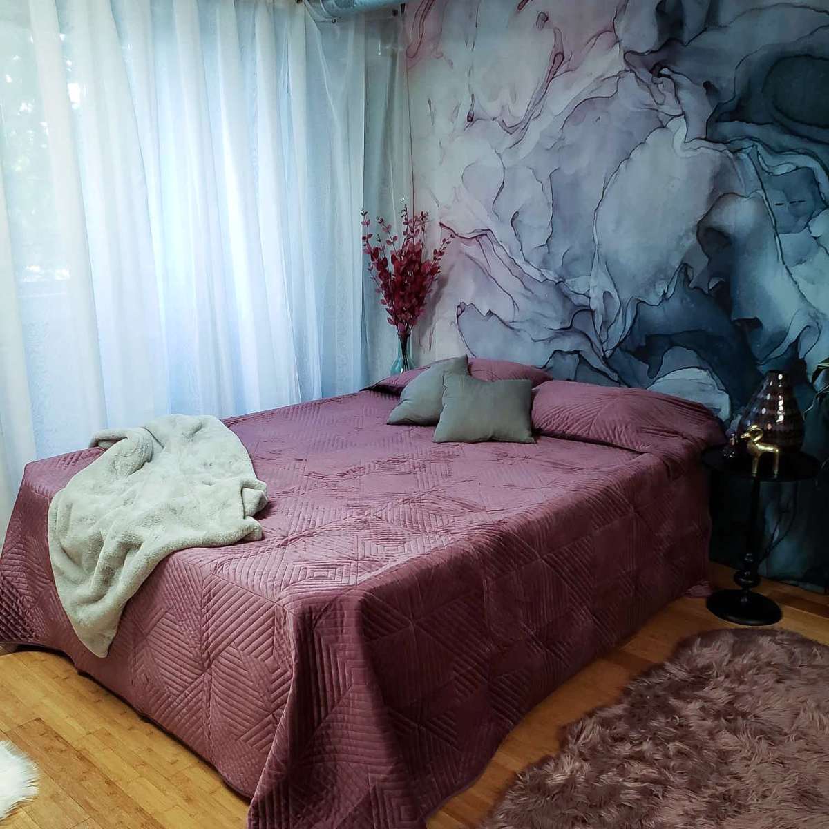 A neatly made bed with a mauve blanket and pillows is positioned against a wall featuring a flower-patterned mural, accompanied by sheer curtains and decorative items.