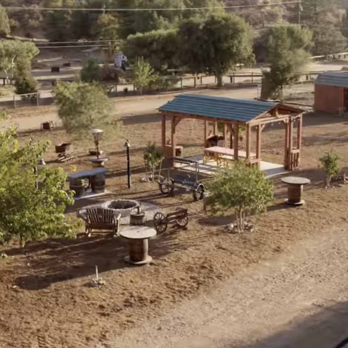 An outdoor area with wooden structures, including a covered gazebo, surrounded by trees and garden furniture.