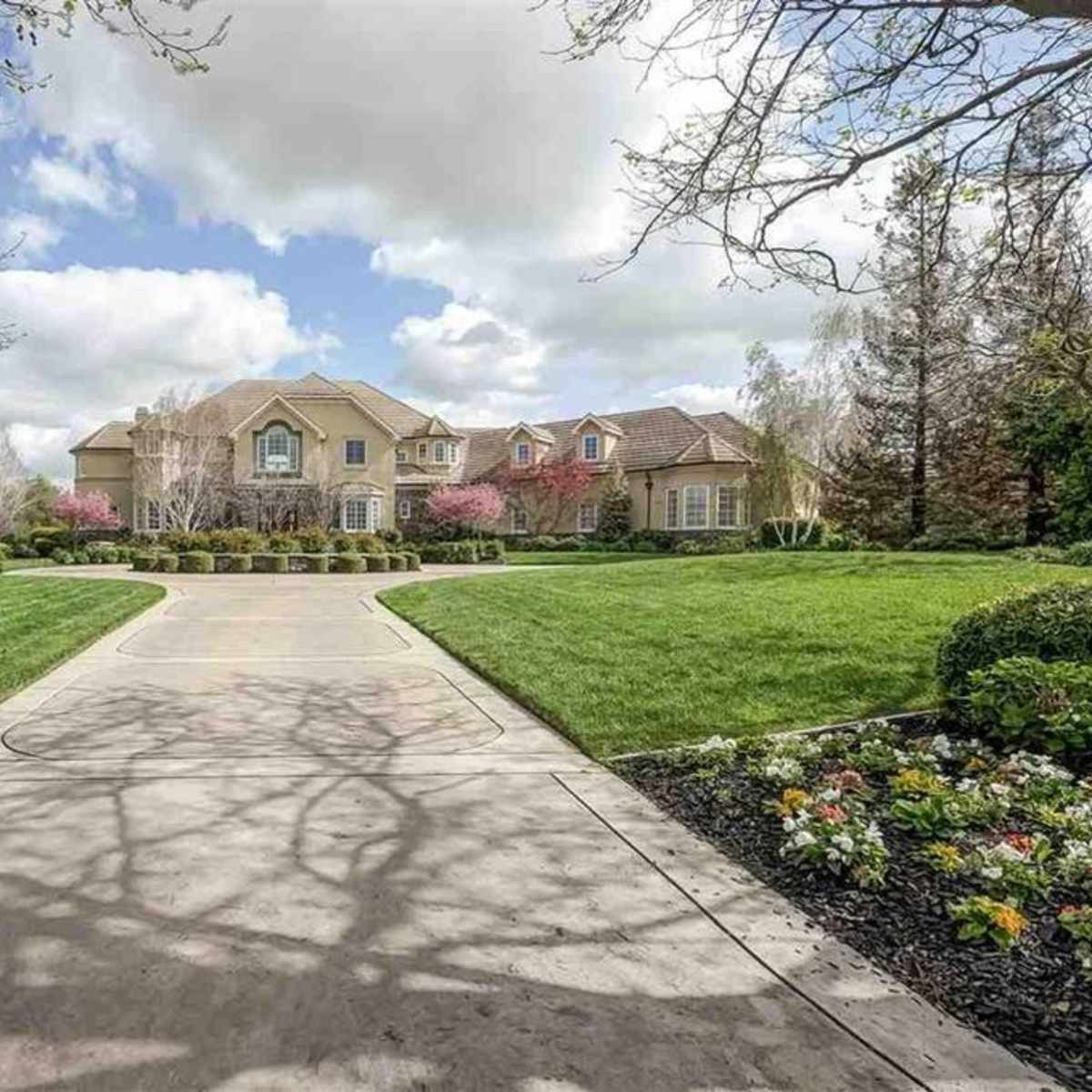 A large, well-manicured house is situated at the end of a wide, curved driveway, surrounded by green lawns and colorful flowerbeds under a cloudy sky.