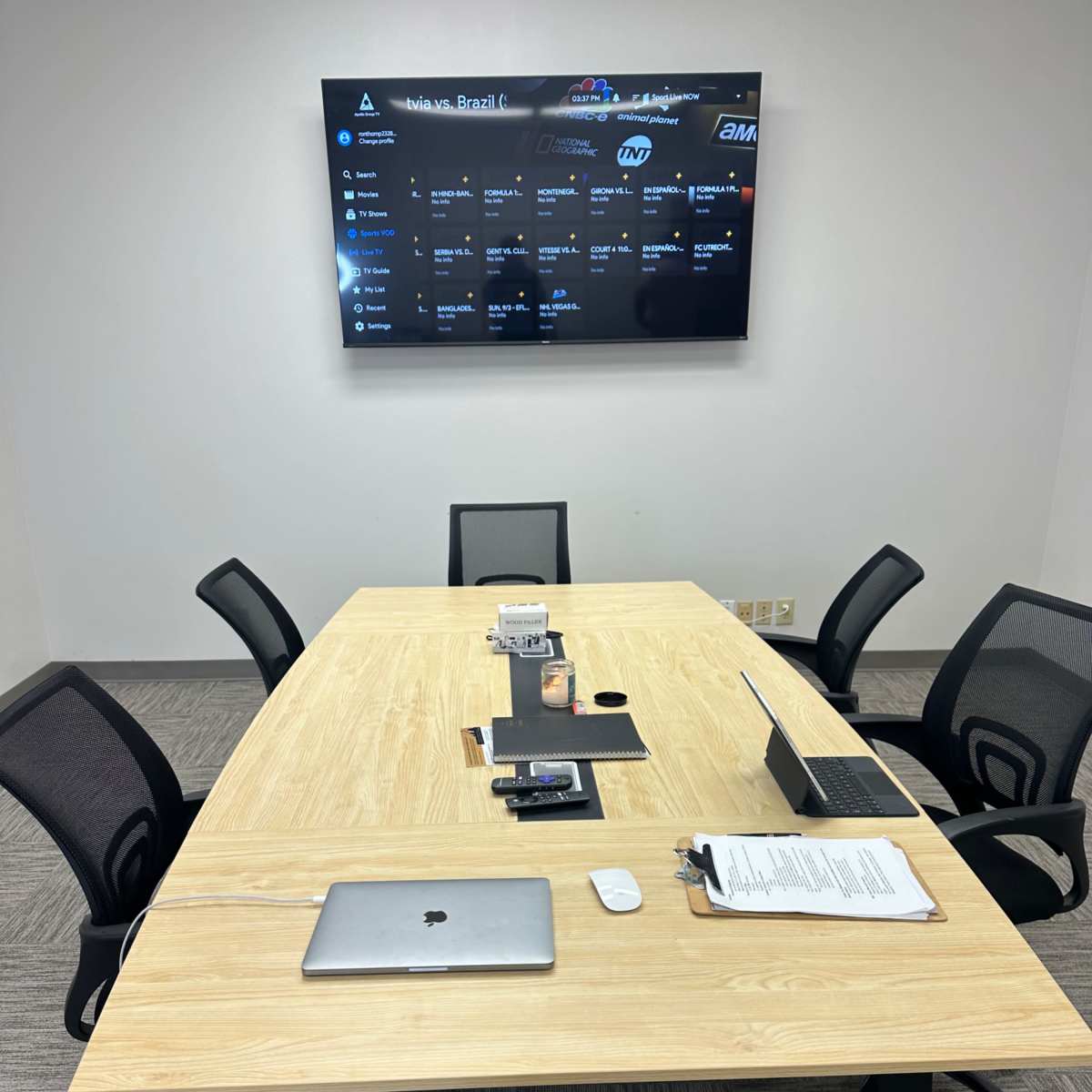 A conference room features a wooden table surrounded by black chairs, with a large screen displaying data on the wall and various electronic devices placed on the table.