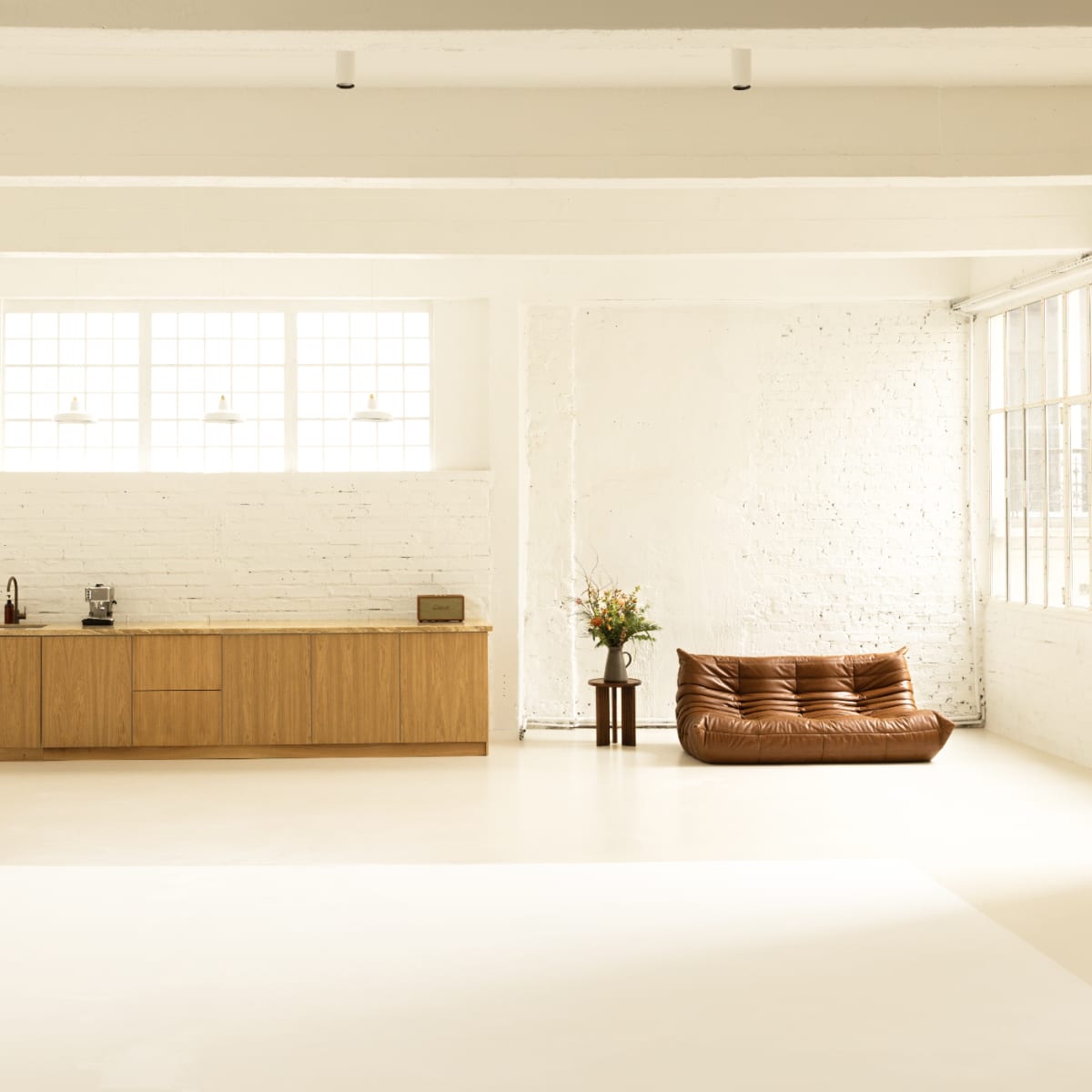 A spacious, minimalist interior with a kitchen area on one side and a brown sofa positioned near large windows on the opposite side.