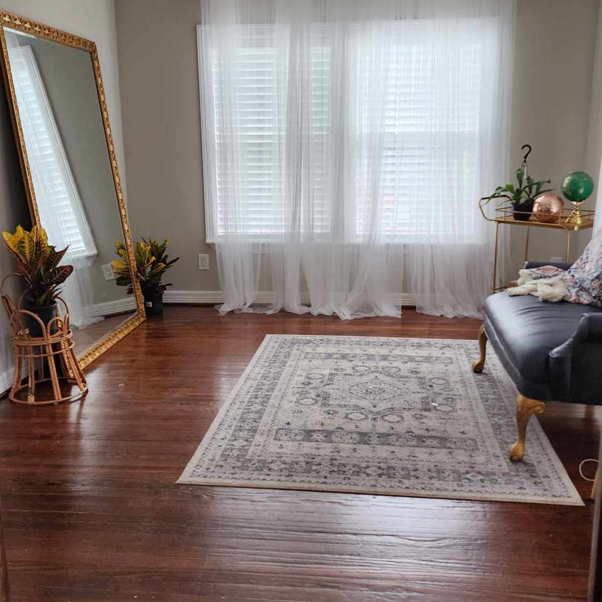A spacious living area features a large mirror, a dark sofa, and a decorative rug on hardwood flooring, with light filtering through sheer curtains.
