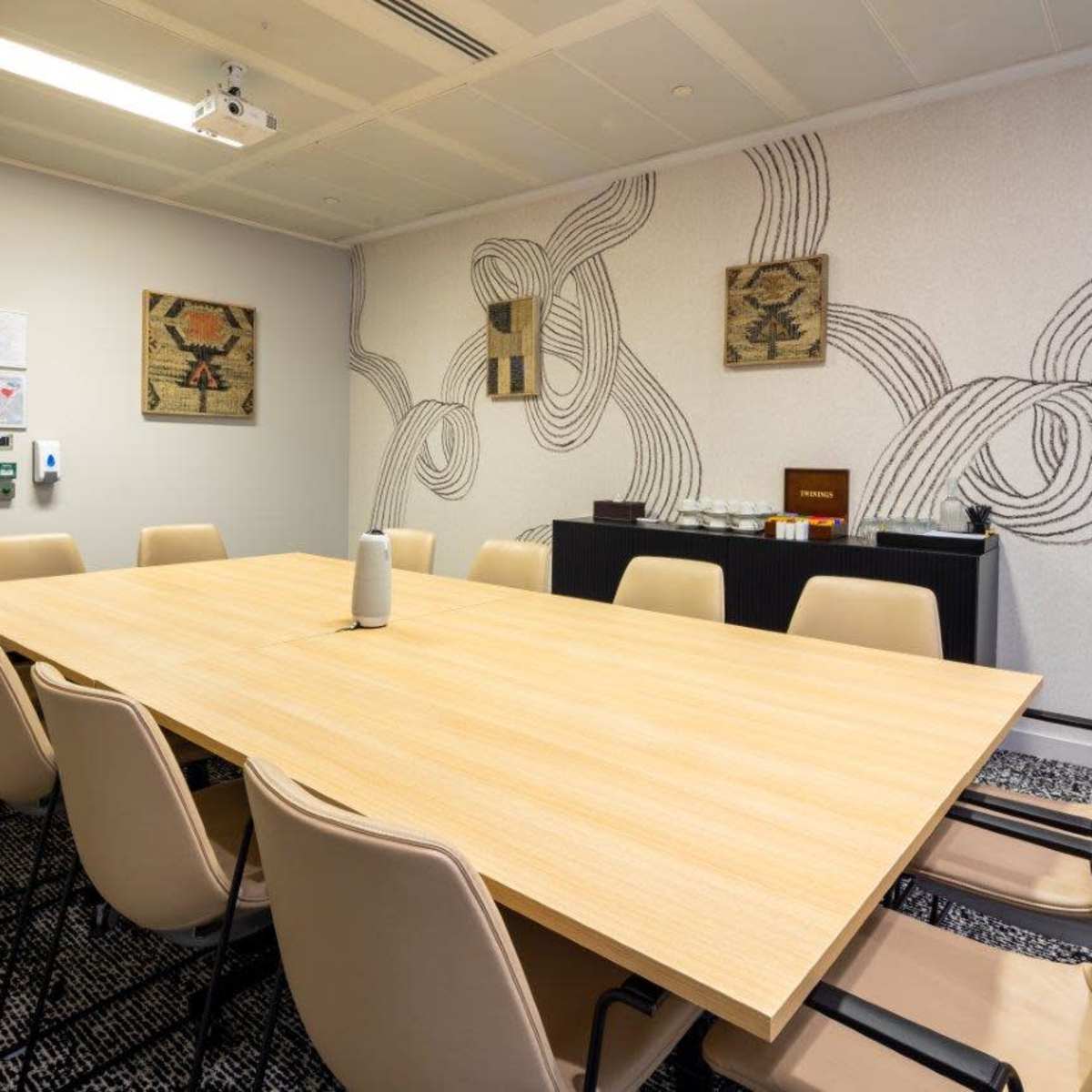 A conference room features a large wooden table surrounded by eight chairs, with patterned walls and a side table displaying refreshments.