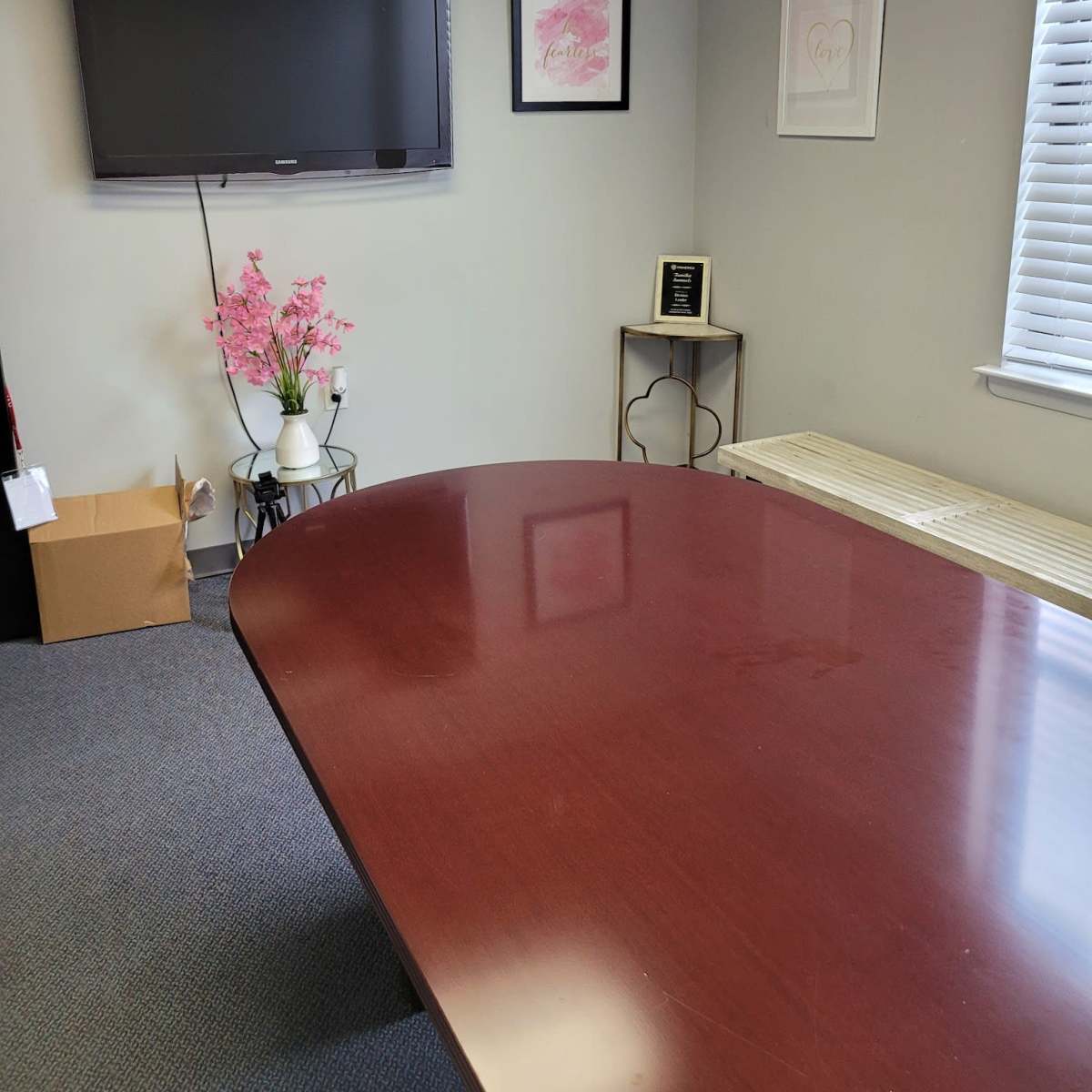 A conference room with a large wooden table, a wall-mounted television, framed artwork, and a decorative plant near the corner.