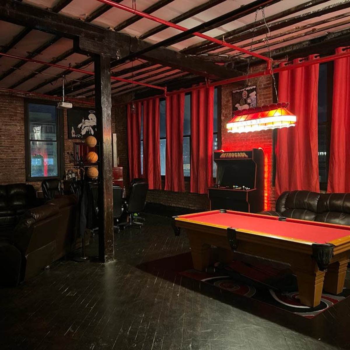 The scene features a dimly lit room with a pool table, leather couches, exposed brick walls, and large red curtains.
