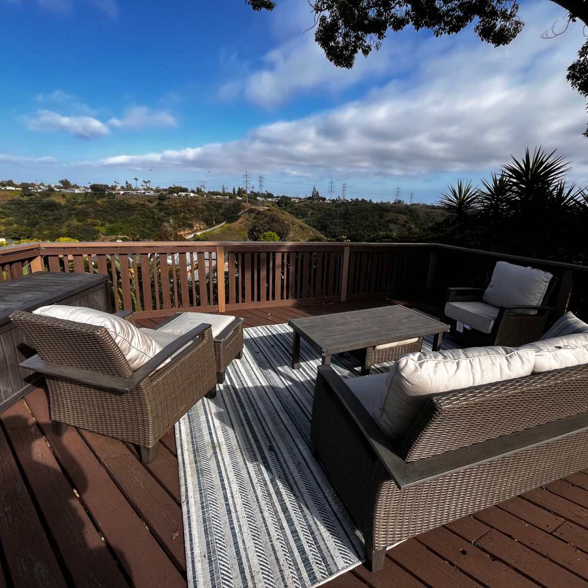 A deck furnished with a seating area that includes two chairs and a table, overlooking a landscape of rolling hills and power lines under a partly cloudy sky.