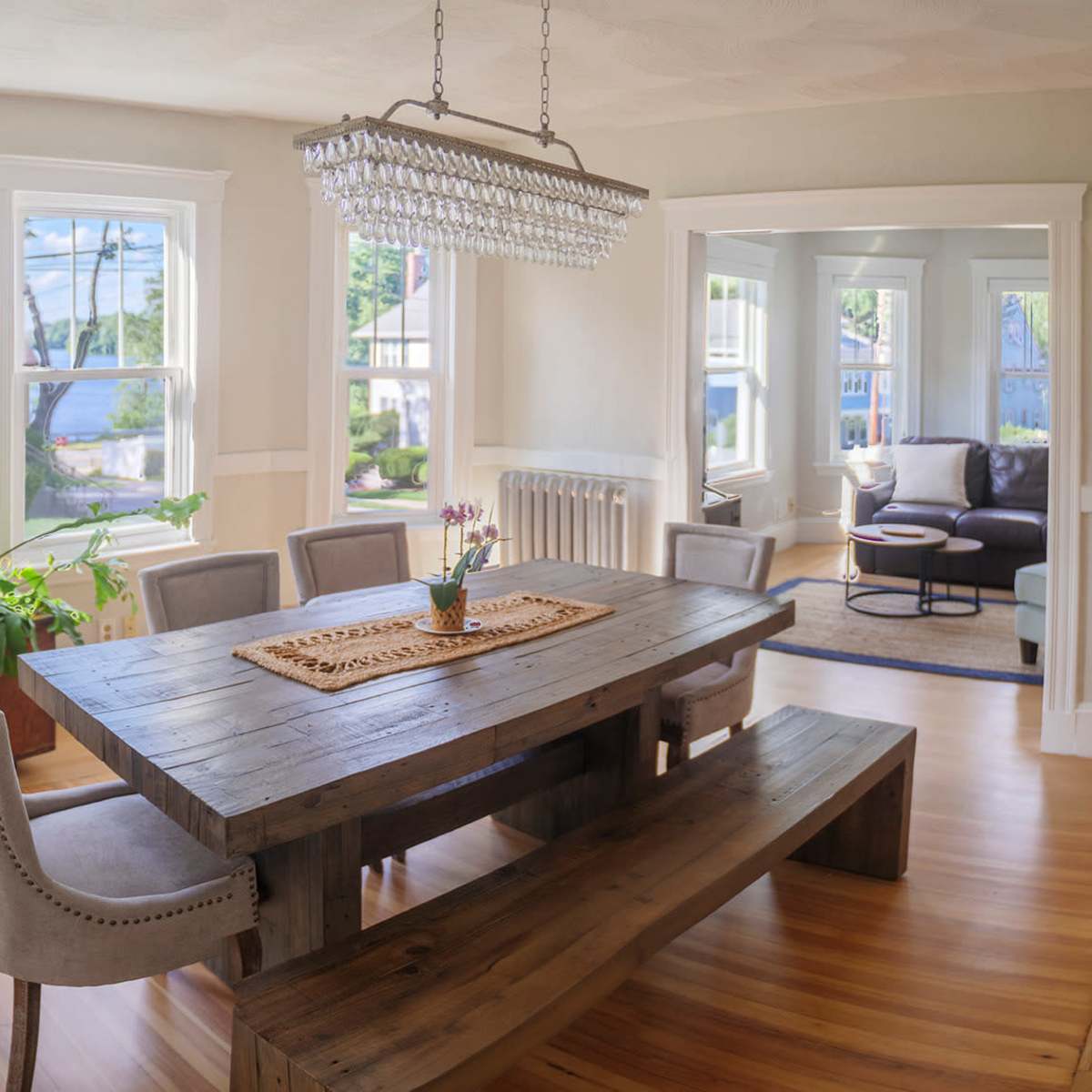 A bright dining area with a wooden table surrounded by upholstered chairs, a crystal chandelier above, and large windows providing natural light.