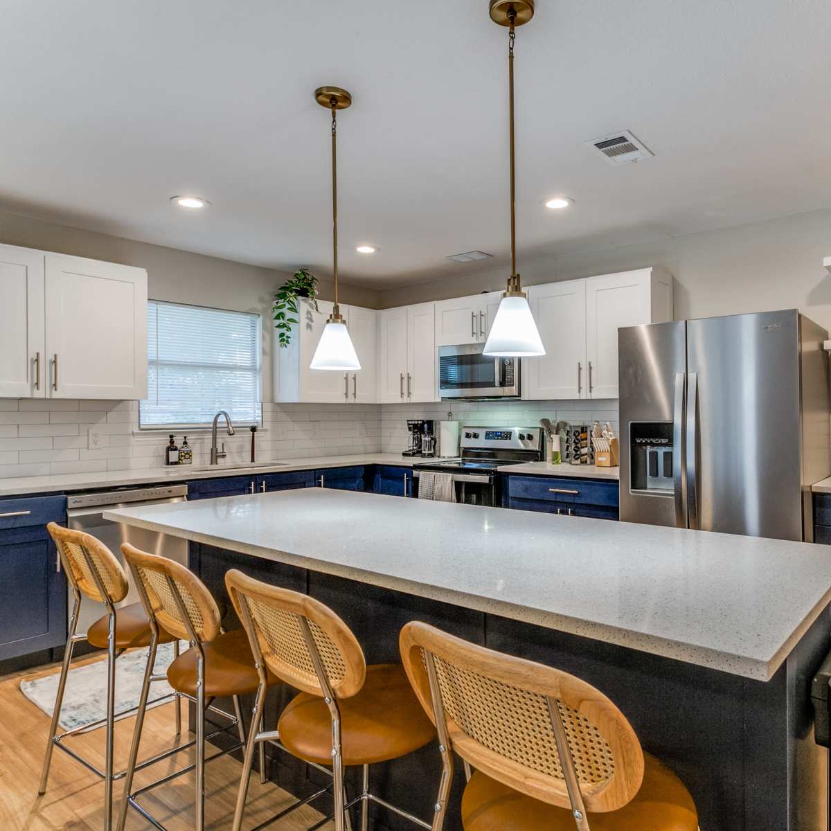 A modern kitchen featuring a central island with four wicker barstools, stainless steel appliances, and white and navy cabinetry.