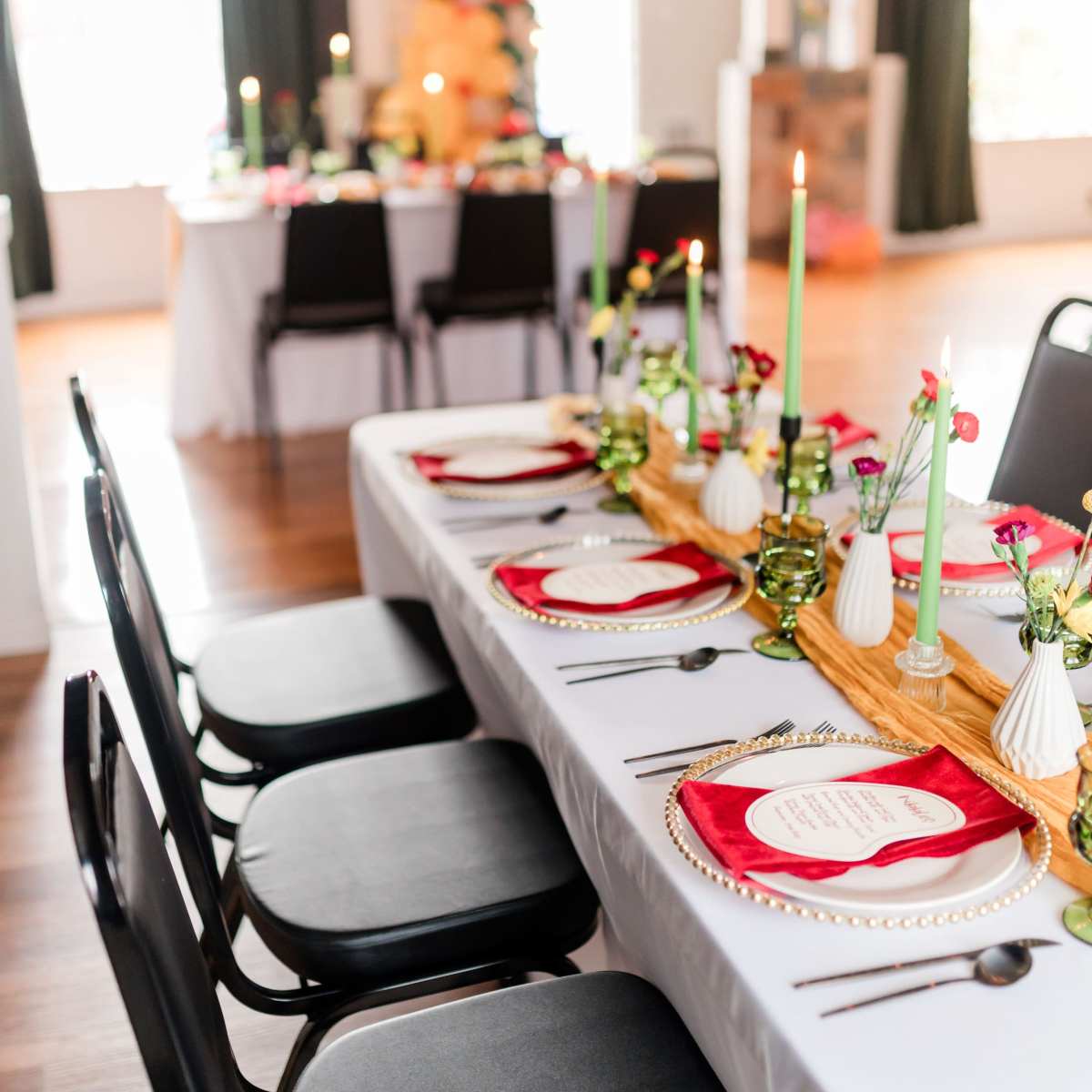 A long dining table is set with red plates, green candles, and decorative centerpieces, while a second table with a festive arrangement is visible in the background.