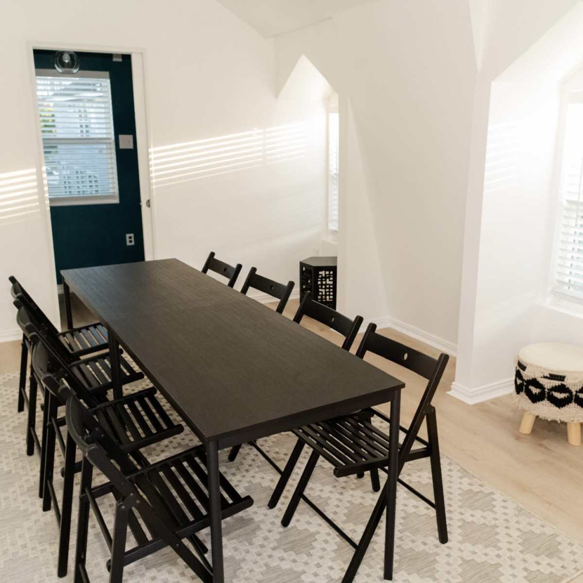 A long black table with eight chairs is set in a bright room with large windows and minimalist decor.