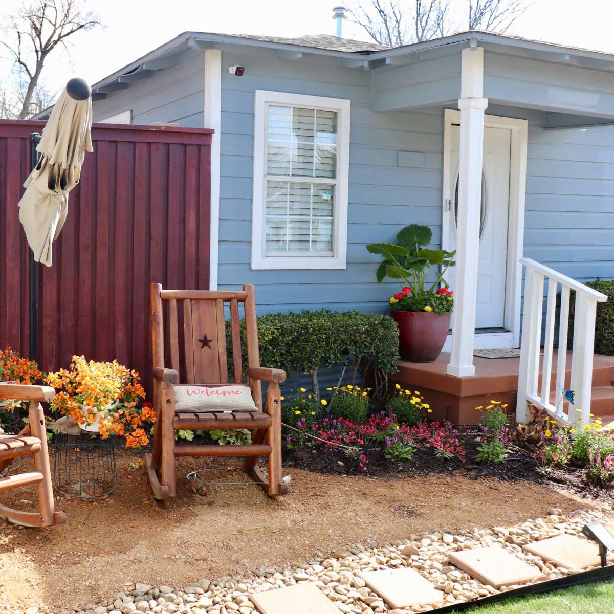 Two rocking chairs with star motifs sit on a gravel path beside a house with blue siding, framed by a wooden fence and colorful flowerbeds.