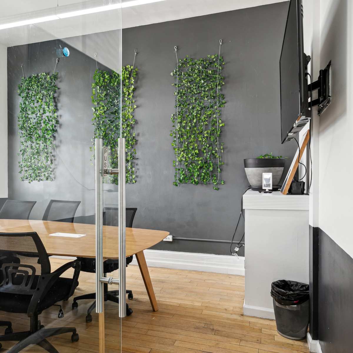 A modern conference room with a wooden table surrounded by black chairs, features green wall installations, and a wall-mounted TV.