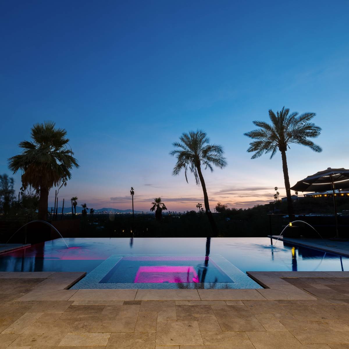 A modern infinity pool with a pink light feature is surrounded by palm trees against a twilight sky.