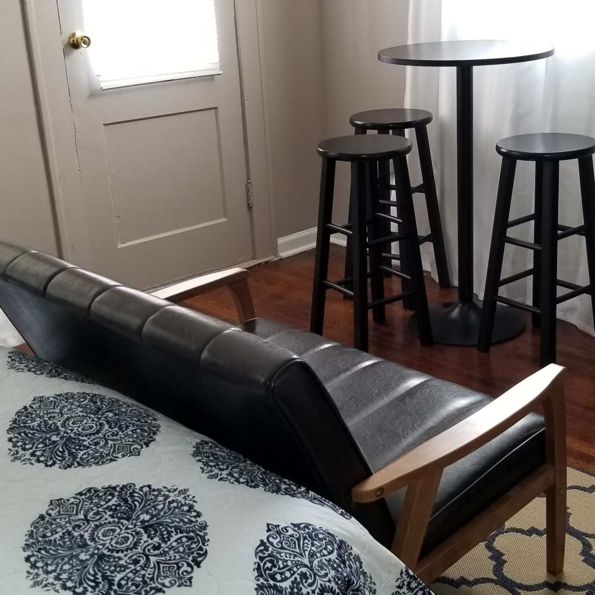A small room with a bed in the foreground and a black table with three stools in the background, next to a chest of drawers.