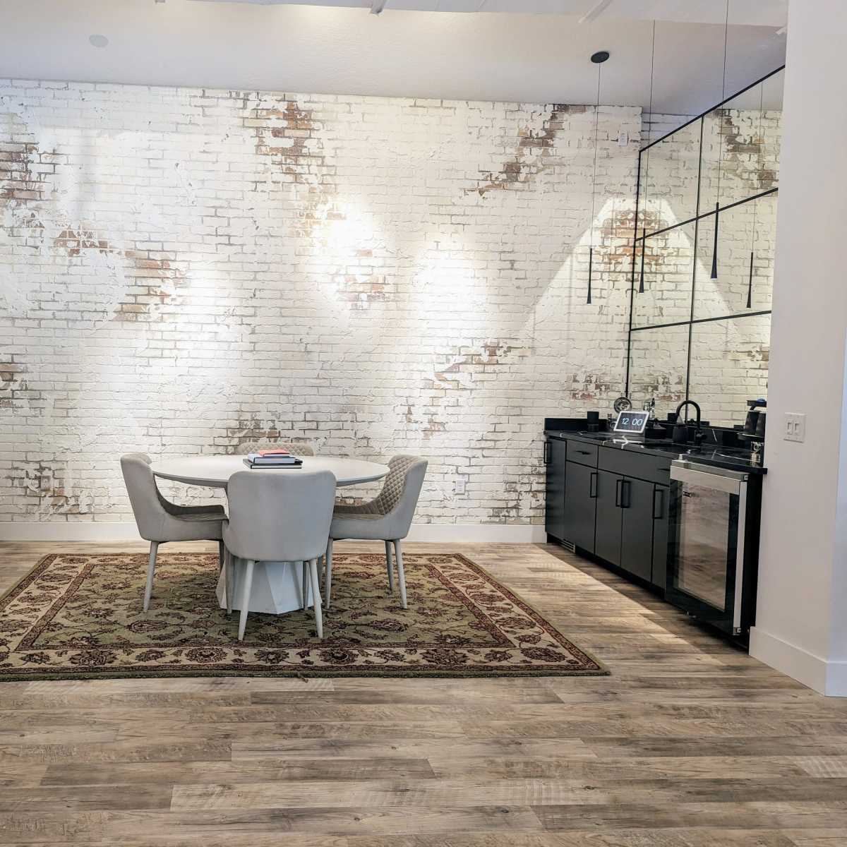 A modern dining area with a round table and four chairs, set against a white brick wall, accompanied by a television and a kitchenette.