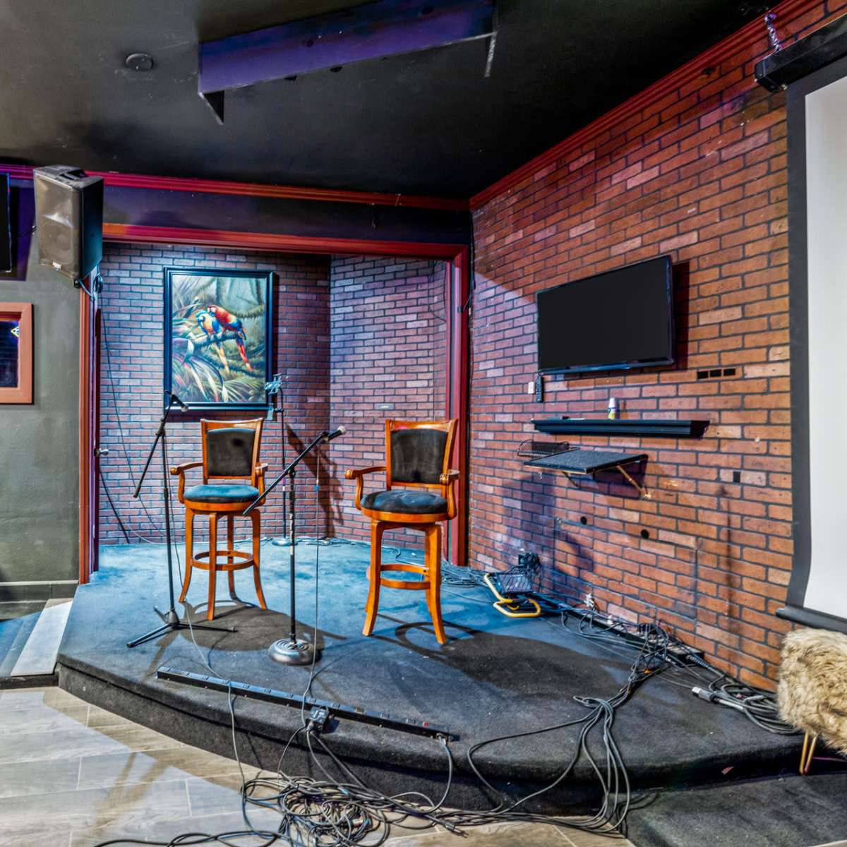 A stage with two empty chairs, microphones, and tangled cables in a dimly lit room featuring brick walls and several screens.