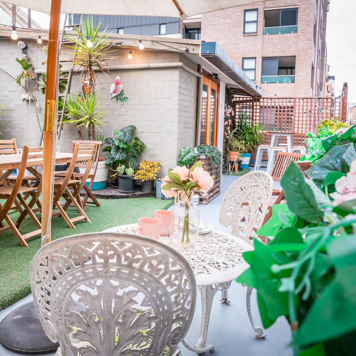 A rooftop garden with wooden dining tables, potted plants, and decorative chairs surrounded by a simple, modern building.