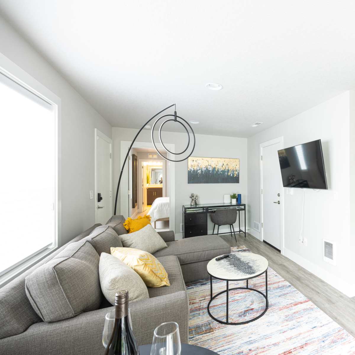 A modern living space featuring a gray sectional sofa, a circular coffee table, and a mounted television, with a small dining area and a bedroom visible in the background.