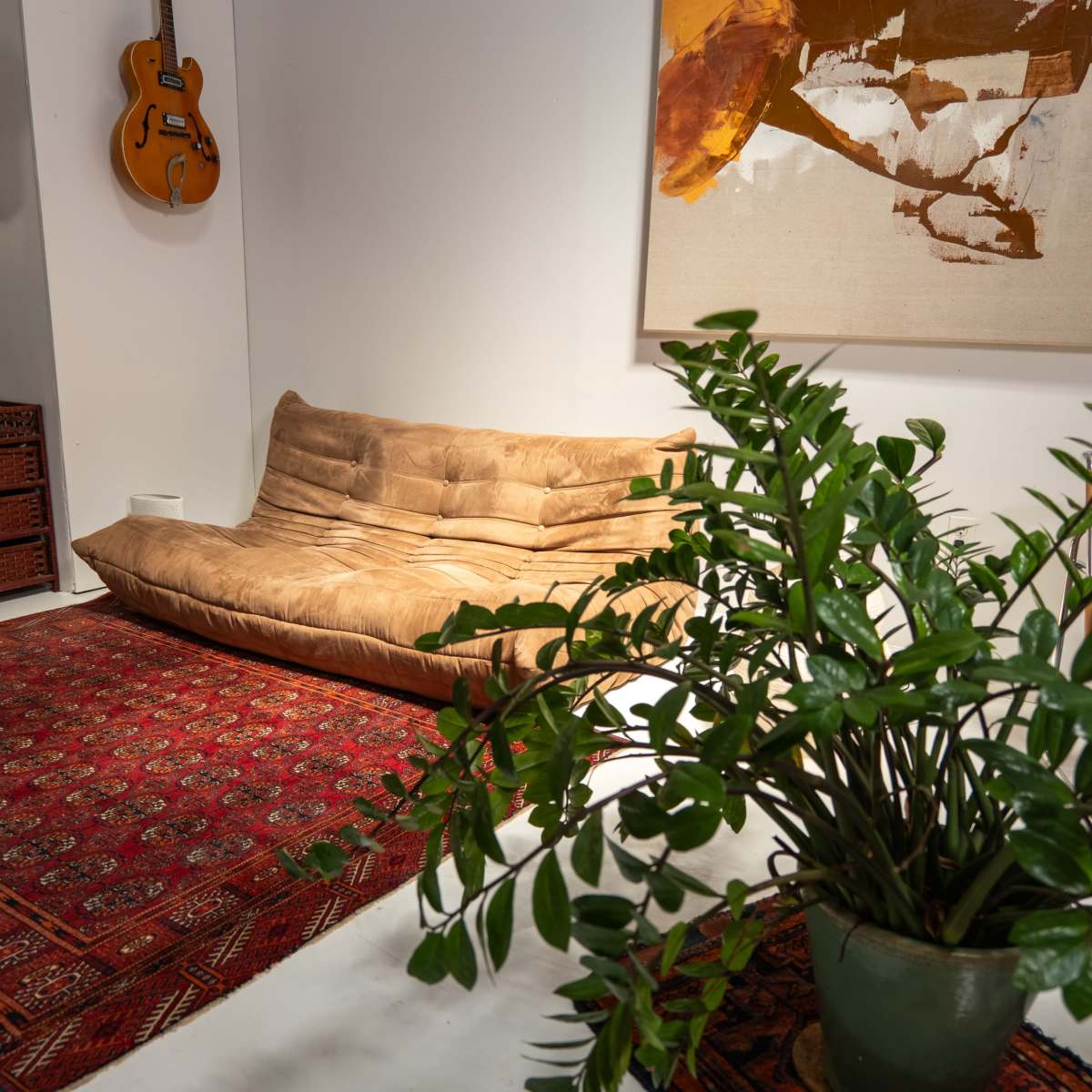 A living space featuring a beige sofa, a decorative plant in a pot, an ornate rug, and a guitar hanging on the wall beside a large abstract painting.
