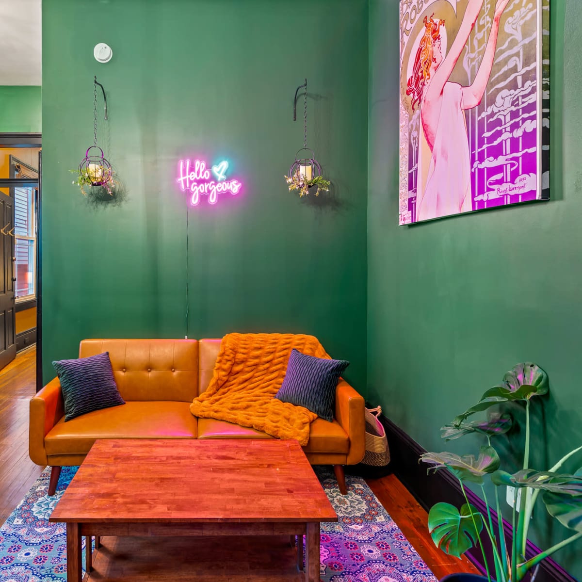 A vibrant room features a brown sofa with a colorful throw, a wooden coffee table, and green walls adorned with plants and a neon sign.