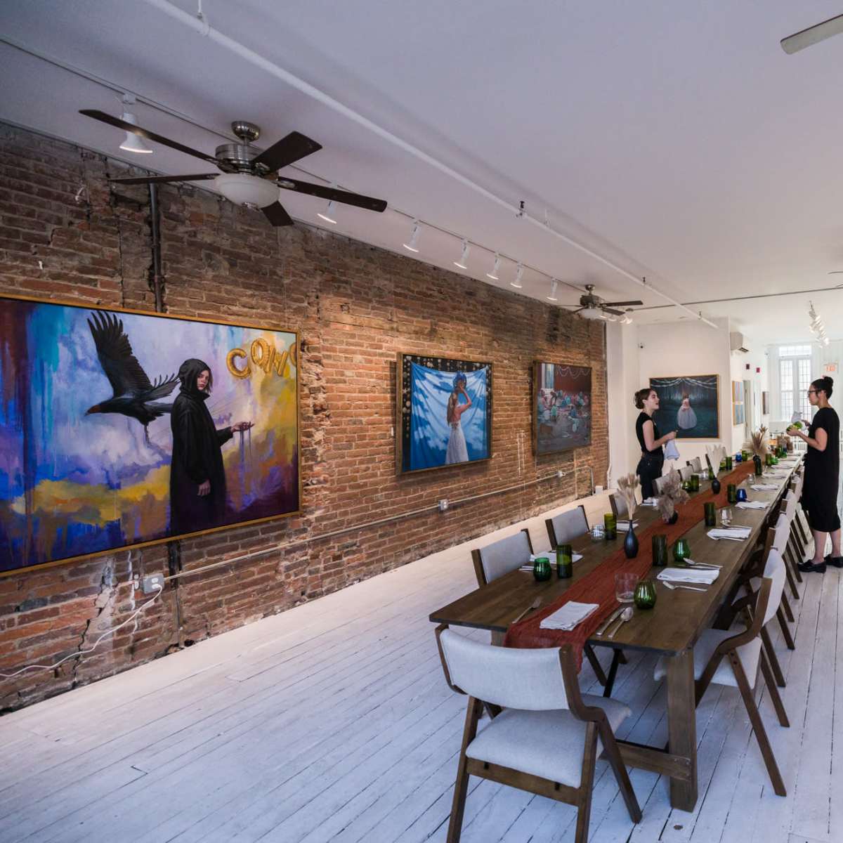 A long wooden dining table is set with greenery in a gallery featuring large paintings on exposed brick walls.
