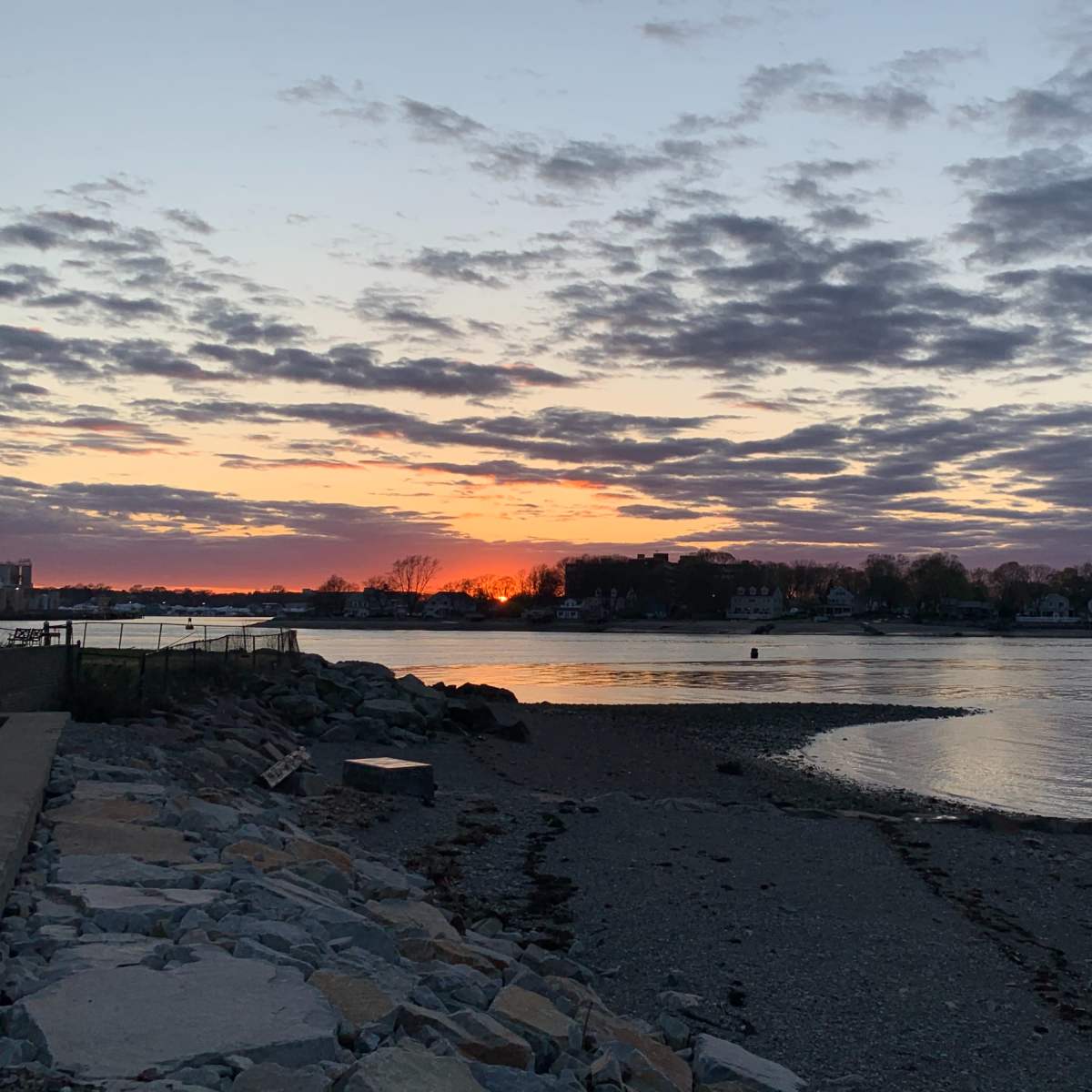 A sunset over a calm body of water, with clouds in the sky and a rocky shore in the foreground.