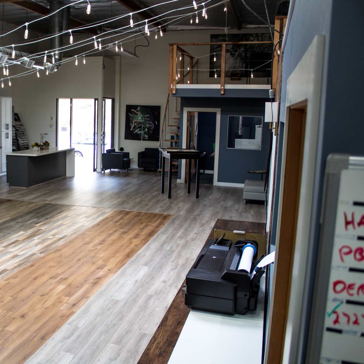 A modern interior space with various flooring samples displayed, a kitchen area, and a seating area lit by decorative string lights.