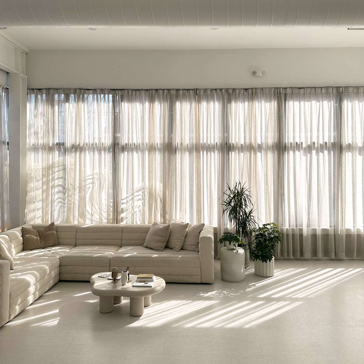 A spacious living room features a light-colored sectional sofa, a round coffee table, and large windows with sheer curtains allowing sunlight to filter in.