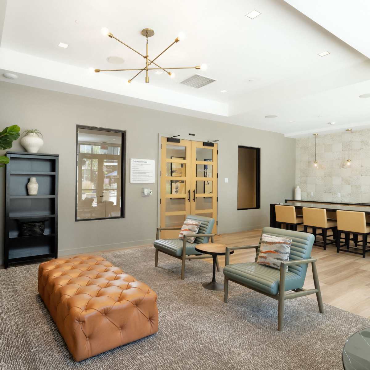 A modern lounge area with a tufted brown ottoman, green chairs, and a kitchenette in a well-lit space with a contemporary design.