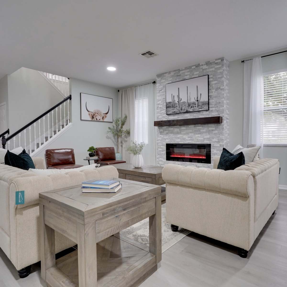 A modern living room features light-colored furniture, a faux stone fireplace, and large windows with sheer drapes.