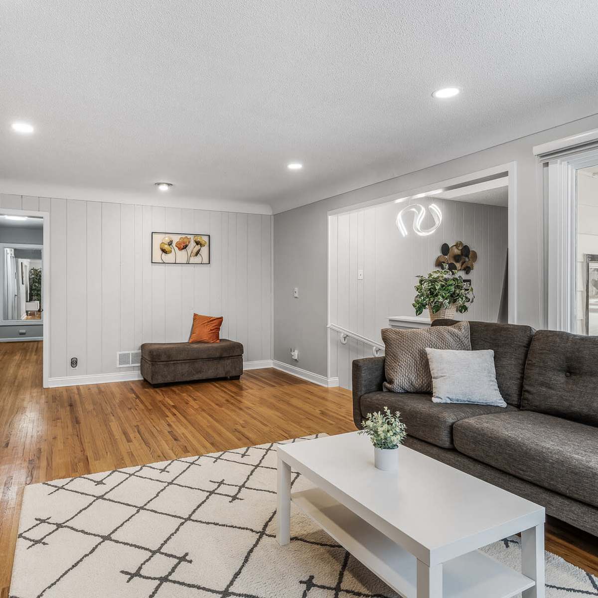The living room features a gray sofa and an orange accent chair, with hardwood floors and white walls complemented by a patterned area rug.