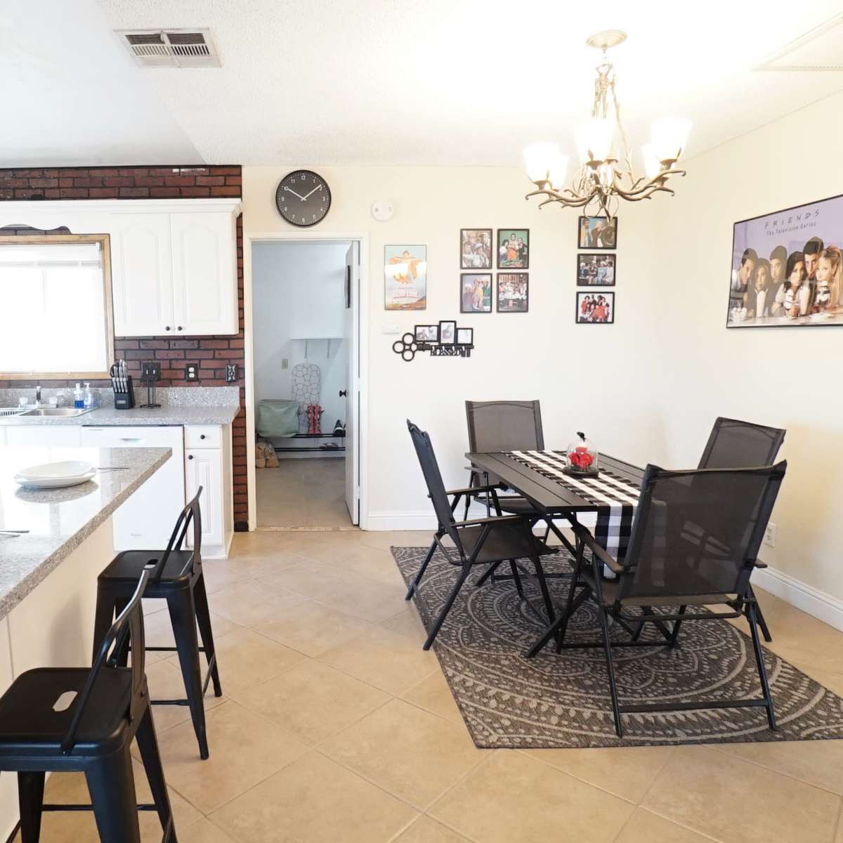 A kitchen and dining area featuring a granite countertop with bar stools, a dining table set for six, and a combination of white cabinets and exposed brick wall.