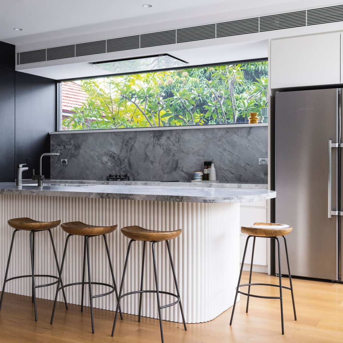 A modern kitchen features a marble-topped island with four wooden stools, stainless steel appliances, and large windows overlooking greenery.