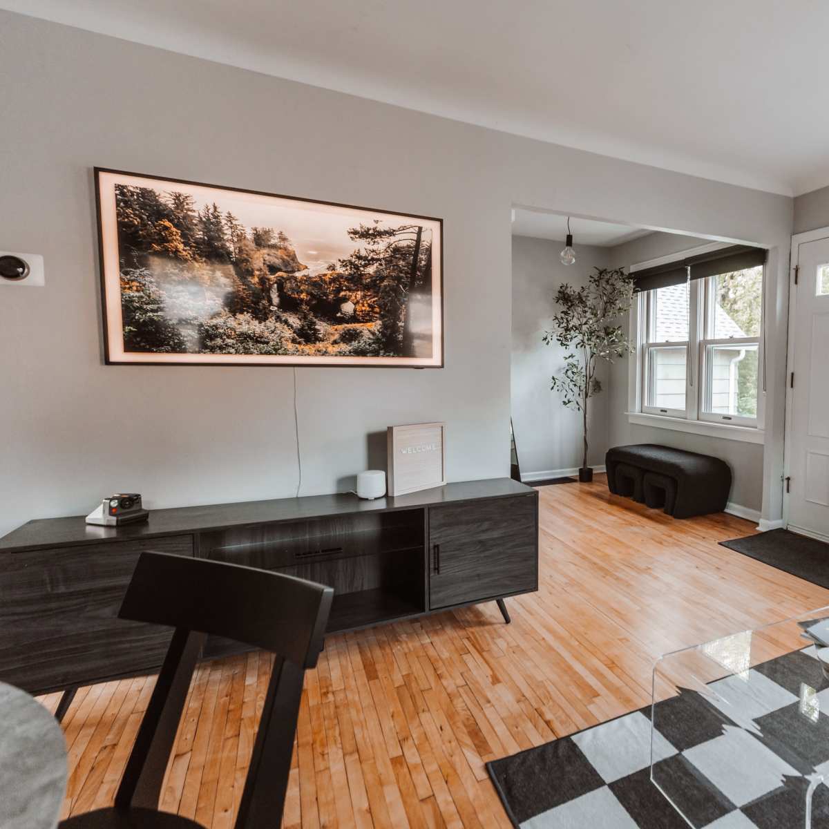 The living room features a wooden floor, a large framed landscape picture on the wall, a sleek black cabinet, a glass coffee table on a checkered rug, and a white armchair.
