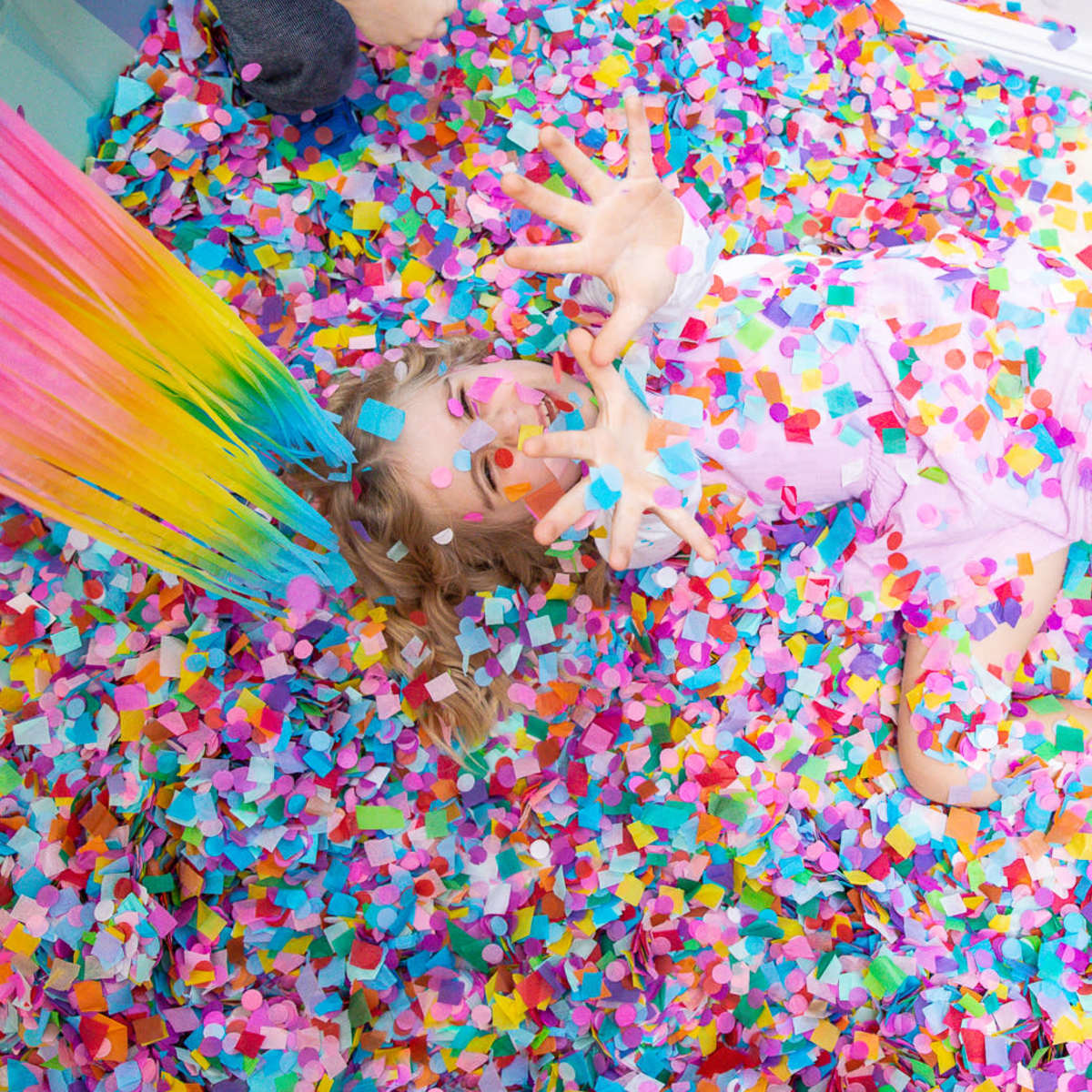 A child lies among a bed of colorful confetti, surrounded by vibrant strands of tissue paper.