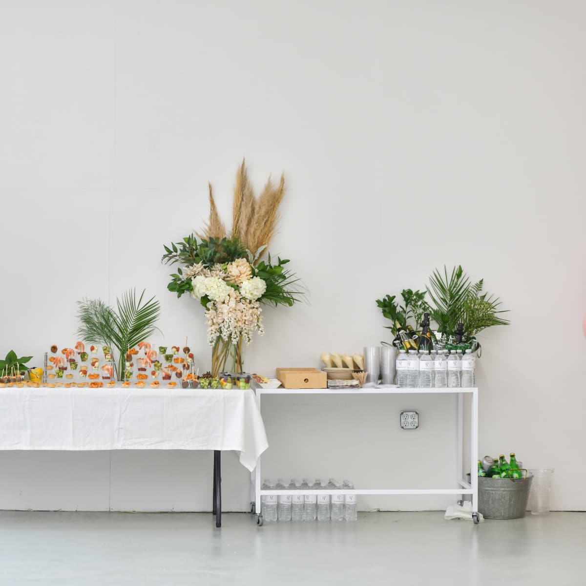 A table with a white cloth is set up against a white wall, displaying an assortment of drinks and snacks, while potted plants add greenery to the scene.