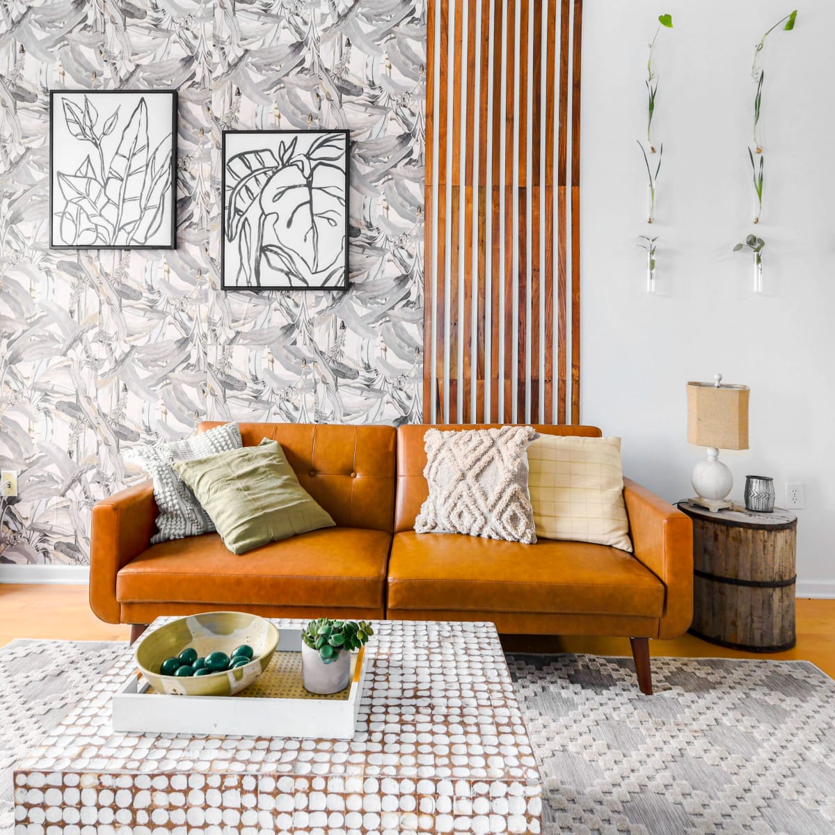 A stylish living room featuring a brown leather sofa, patterned wallpaper, two framed artworks, a coffee table with decorative items, and a wooden side table.