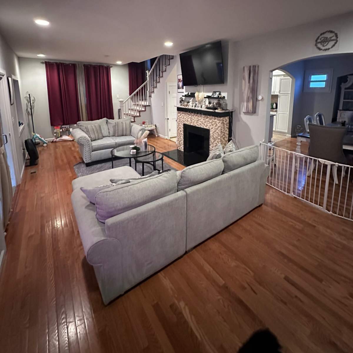 A spacious living room with gray sofas, a fireplace, a large television mounted on the wall, and a staircase leading upstairs.